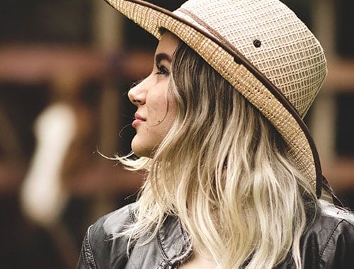 Girl with light balayage hair with slight wave looking to the side wearing a wide natural brimmed hat and a dark brown leather jacket