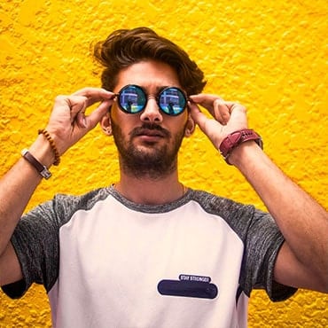Guy holding his hands up to his reflective mirror sunglasses in front of a bright yellow wall