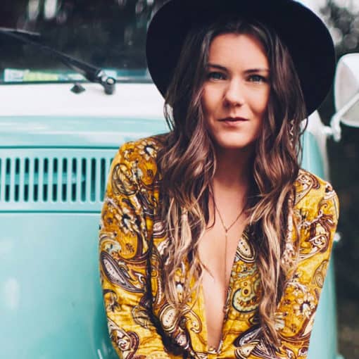 Brunette girl with hat and bright paisley shirt standing in front of vintage car