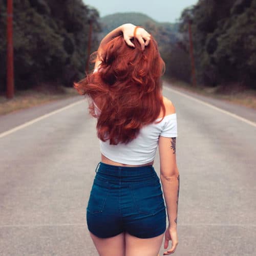 bright red head lady in centre of country road mountains in background wearing denim shorts and whitetop with some visible tattoos on right arm