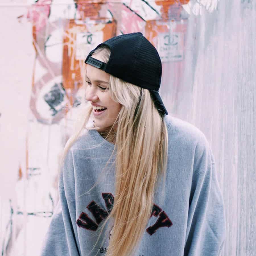 Girl with long blonde hair, wearing a cap on backwards, standing in front of a graffiti wall