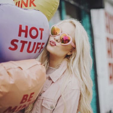 Girl with blonde hair, pink love heart sunglasses on, holding candy coloured love heart shaped balloons. They say, hot stuff and be mine. She is pretending to blow a kiss