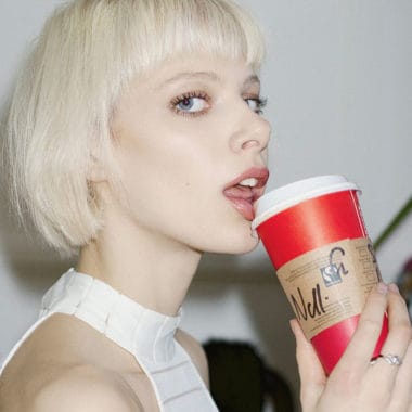 Bowl cut that is bleached blonde. Girl sipping on a large coffee cup.