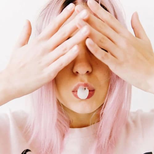 lady with soft pink long bob with her hands over her face and has just blown a chewing gum bubble