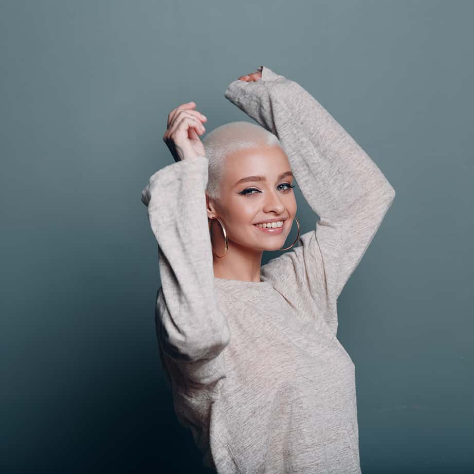 Lady with platinum blonde buzz cut and large hoop earrings holding her arms up in the air