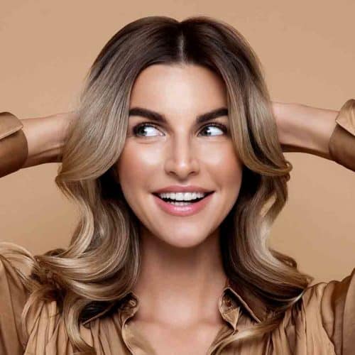 Female with creamy blonde, wavy hair, wearing a bronze blouse, holding both hands behind head.