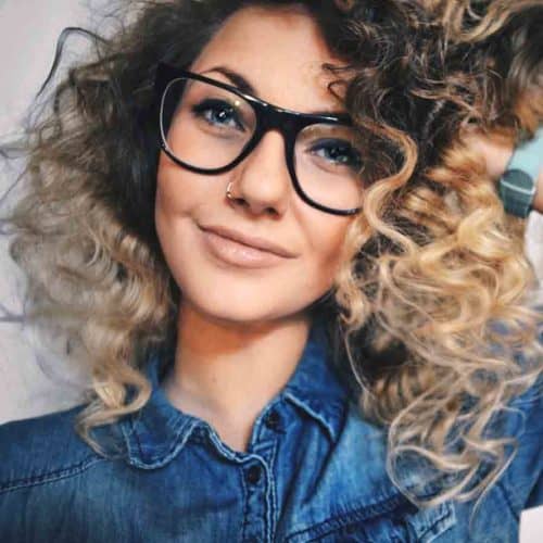 Female with medium curly hair, dark roots and light ends. Wearing large black glasses and hand pushing side of head.