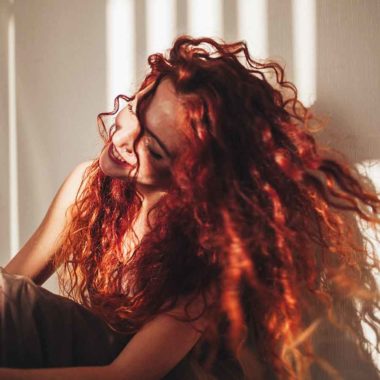 Long, curly ,red-head female in room with dramatic sunshine across face, looking away, swishing hair.