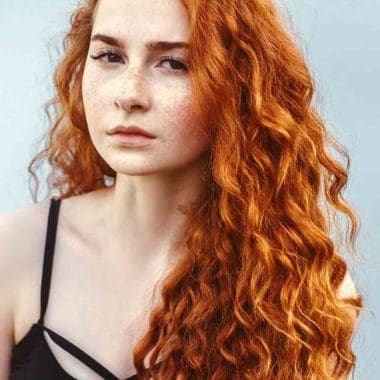 Long, curly, red head female with freckles, standing against white wall