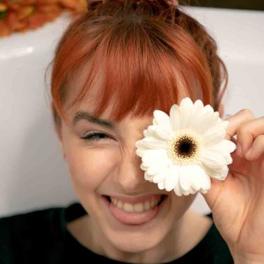 Face of copper hair female sitting in bath-tub, holding a white flower in front of her left eye while biting on tongue.