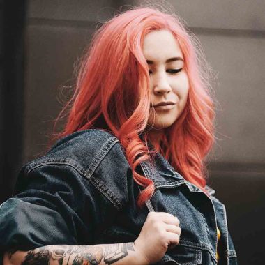 Side view of female with long peach, curled hair, wearing denim jacket, holding handbag strap over shoulder..